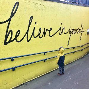 Person stands in front of a yellow painted wall where it reads "believe in yourself"